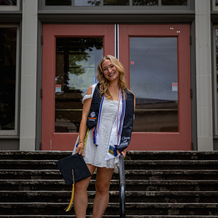 Katie smiles and poses in a graduation stole and cords, holding a grad cap.