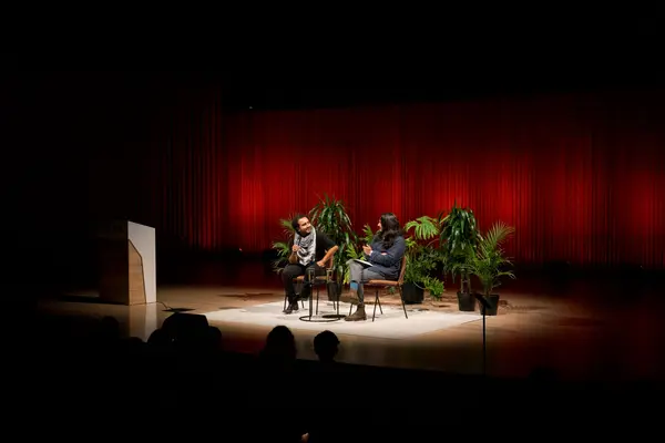 Javier and Dr. Zapata sitting on stage in front of a red curtain and plants, talking into mics