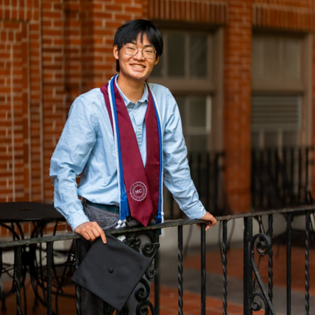 Kevin smiles and poses in an HC stole and cords with a grad cap in hand.