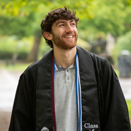 Matthias smiles and looks away from the camera into the distance, wearing a graduation gown, cords, and HC stole.
