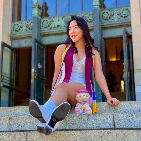Sophiana sits on the MU stairs, smiling and posing in an HC stole and cords with a grad teddy bear nearby.